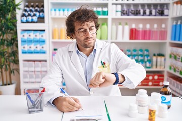 Hispanic young man working at pharmacy drugstore checking the time on wrist watch, relaxed and...