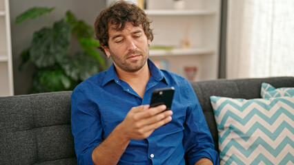 Young hispanic man using smartphone sitting on sofa at home