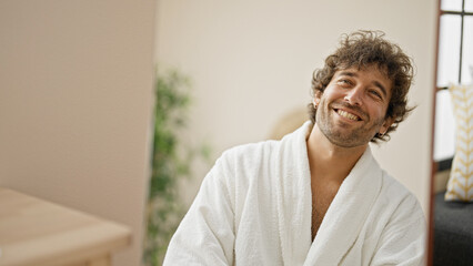 Young hispanic man wearing bathrobe looking on mirror smiling at home