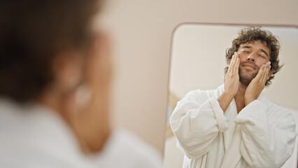 Young hispanic man wearing bathrobe applying skin face lotion looking on mirror at home