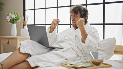 Young hispanic man wearing bathrobe using laptop celebrating at bedroom