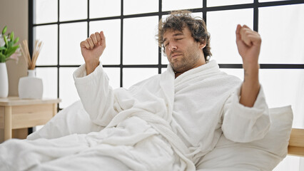 Young hispanic man waking up stretching arms wearing bathrobe at bedroom