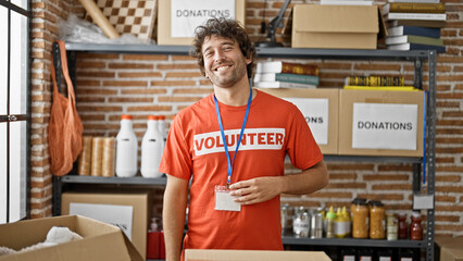 Young hispanic man volunteer smiling confident standing by donations cardboard box at charity center