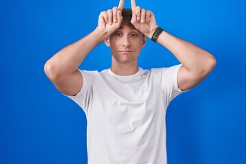 Naklejka premium Caucasian blond man standing over blue background doing funny gesture with finger over head as bull horns