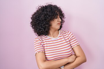 Young middle east woman standing over pink background looking to the side with arms crossed convinced and confident