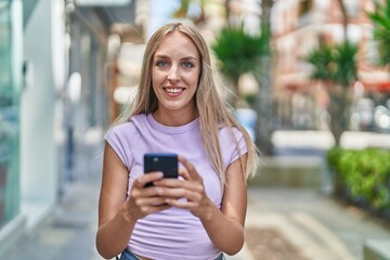 Young blonde woman smiling confident using smartphone at street