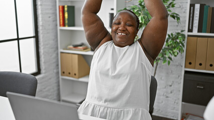 Confident plus size african american woman, a successful office worker, using laptop and celebrating a win with arms raised at her desk indoors