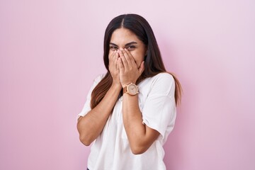 Young arab woman standing over pink background laughing and embarrassed giggle covering mouth with...