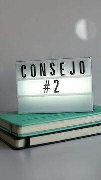 Vertical. Letrero retro-iluminado con el texto "Consejo 2" sobre dos cuadernos uno negro y otro turquesa sobre mesa y fondo blanco. Terminando la vista sobre un bote de bol&iacute;grafos. Concepto ideas