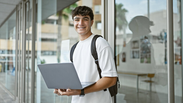 Cheerful hispanic teenager at university, a young, smart student enjoying laptop studies in the sunlight, backpack slung over a casual outfit.