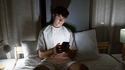 Young hispanic teenager using smartphone sitting on bed at bedroom