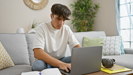 Concentrated young hispanic male student, engrossed in online education, sitting on cozy living...