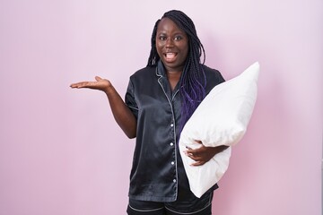 Young african woman wearing pijama hugging pillow pointing aside with hands open palms showing copy...