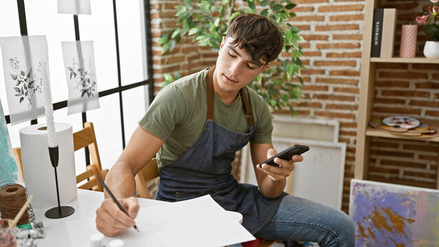 Inside the art studio, passionate young hispanic teenager draws on paper, engrossed in artistry and learning, while casually looking at smartphone amidst a creative buzz