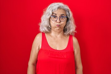 Middle age woman with grey hair standing over red background puffing cheeks with funny face. mouth inflated with air, crazy expression. - obrazy, fototapety, plakaty