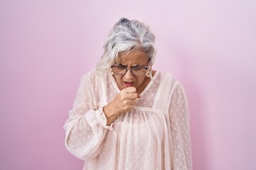 Middle age woman with grey hair standing over pink background feeling unwell and coughing as...