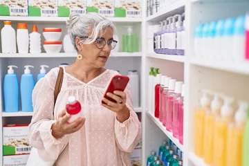 Poster Middle age grey-haired woman customer using smartphone holding medicine bottle at pharmacy © Krakenimages.com