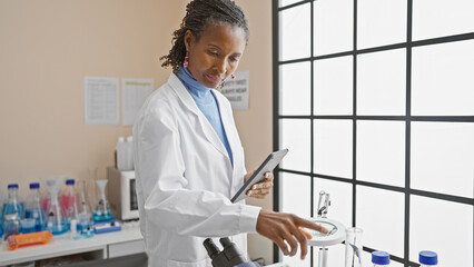 African american woman scientist analyzing samples in laboratory using tablet and microscope