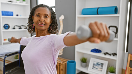 African american woman exercising with dumbbells in a rehab clinic room, portraying health and...