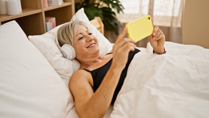 Smiling mature hispanic woman with grey hair lying in bed using smartphone and headphones indoors.