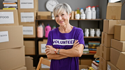 Mature woman volunteer with grey hair smiling in warehouse surrounded by donation boxes.