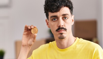 Young caucasian man holding bitcoin with serious expression at new home