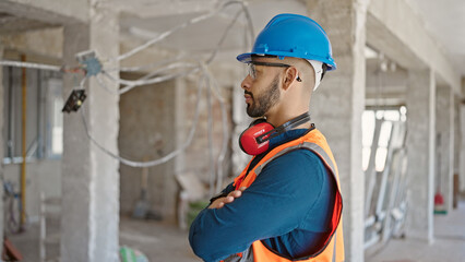 Young hispanic man builder with crossed arms wearing hardhat at construction site