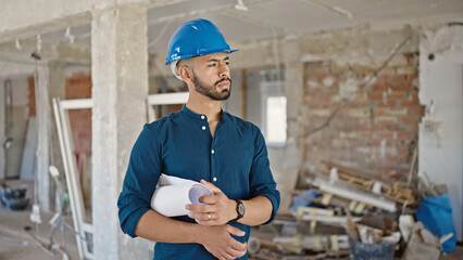 Young hispanic man architect wearing hardhat holding blueprints at construction site