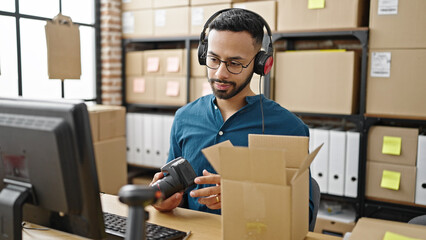 Young hispanic man ecommerce business worker packaging cardboard box wearing headphones at office