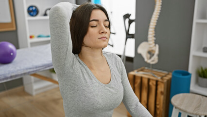Young hispanic woman in physical therapy clinic performing shoulder stretch exercise indoors