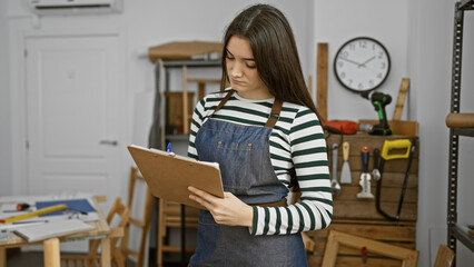 A focused brunette teenager wearing a striped shirt and denim apron stands in a carpentry workshop studying her clipboard.