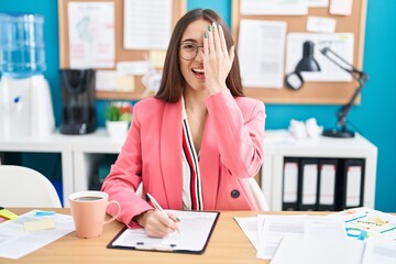 Young hispanic woman working at the office wearing glasses covering one eye with hand, confident smile on face and surprise emotion.