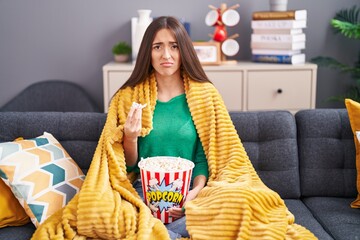 Young hispanic woman eating snack sitting on the sofa at home depressed and worry for distress,...