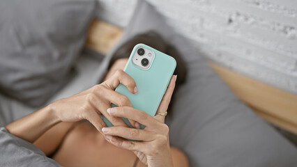 Morning bliss, young beautiful hispanic woman relaxing in bedroom, comfortably lying on bed using smartphone, typing messages online