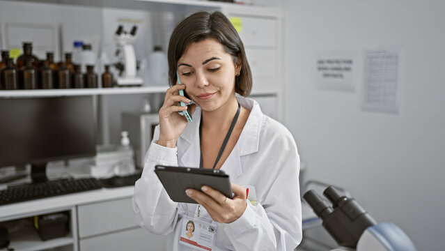 Young beautiful hispanic woman scientist using touchpad talking on smartphone at laboratory