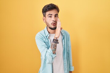 Young hispanic man with tattoos standing over yellow background hand on mouth telling secret rumor,...