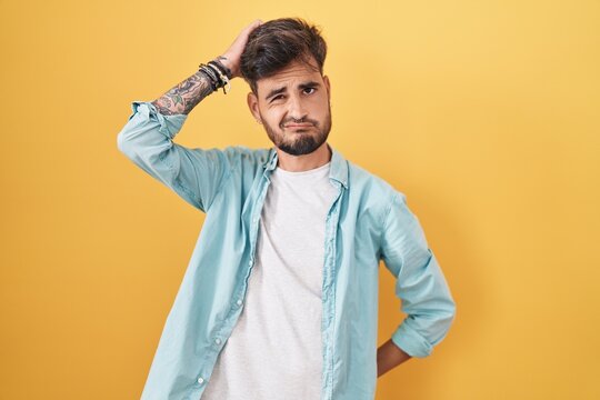 Young hispanic man with tattoos standing over yellow background confuse and wondering about question. uncertain with doubt, thinking with hand on head. pensive concept.