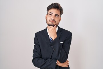 Young hispanic man with tattoos wearing business suit and tie with hand on chin thinking about...