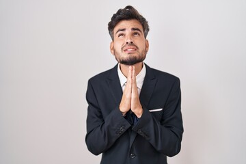 Young hispanic man with tattoos wearing business suit and tie begging and praying with hands...