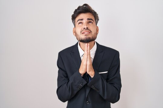 Young hispanic man with tattoos wearing business suit and tie begging and praying with hands together with hope expression on face very emotional and worried. begging.