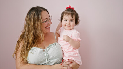 Confident mother joyfully holding her cute baby daughter in her arms, standing against an isolated pink background, radiating positivity and happiness while casually hugging her child.