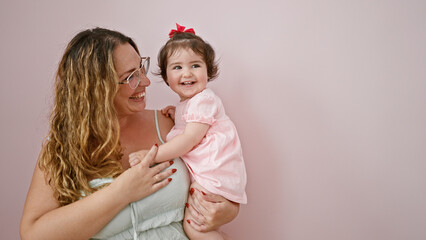 Confident mother joyfully holding her cute baby daughter in her arms, standing against an isolated pink background, radiating positivity and happiness while casually hugging her child.