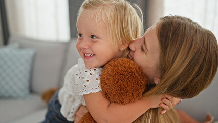 Happy caucasian mother and daughter comfortingly hugging, holding a monkey doll, sitting relaxingly together at their cozy home