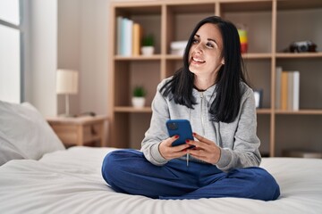 Young caucasian woman using smartphone sitting on bed at bedroom