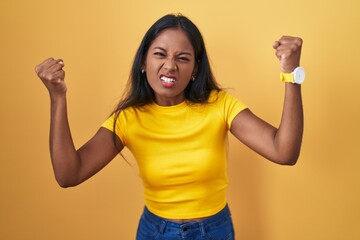 Young indian woman standing over yellow background angry and mad raising fists frustrated and...