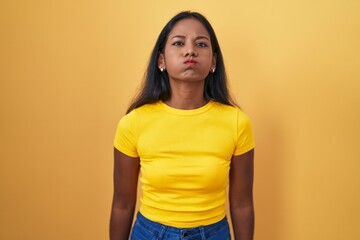 Obraz premium Young indian woman standing over yellow background puffing cheeks with funny face. mouth inflated with air, crazy expression.