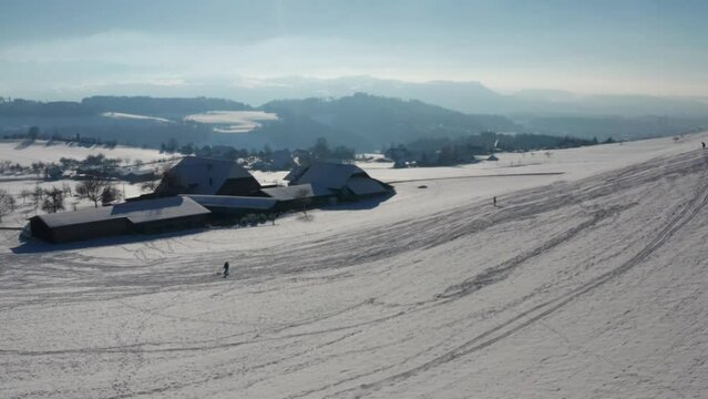 Perfect world in winter: A snowy farm, kids sledding in front in the snow, with the swiss alps in the background. Aerial drone footage in central europea