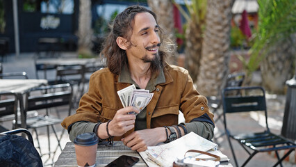 Young hispanic man tourist counting dollars sitting on table at coffee shop terrace