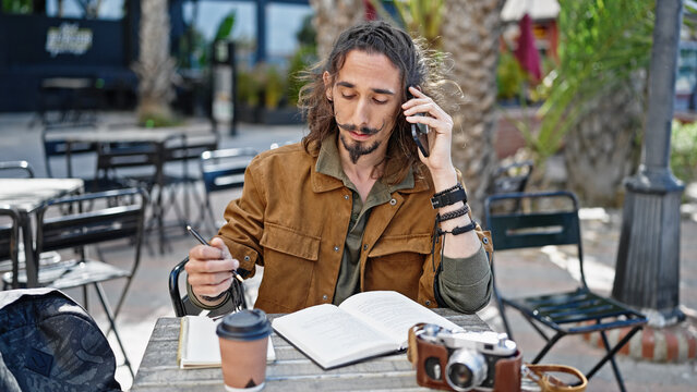 Young hispanic man tourist talking on smartphone sitting on table taking notes at coffee shop terrace