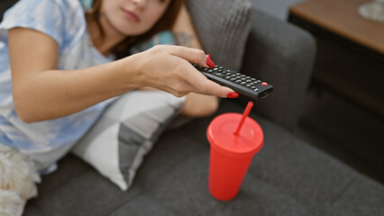 A young woman lounges casually on a sofa in an apartment holding a remote control with a red cup on the side table.